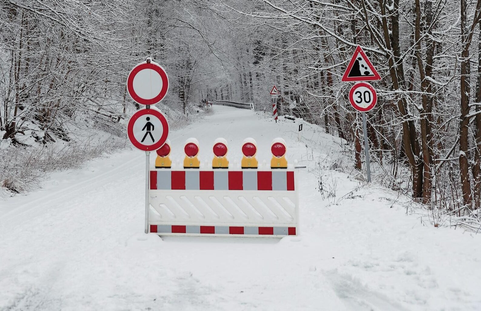 A snow-covered road flanked by trees with warning signs indicating a path closure in winter.