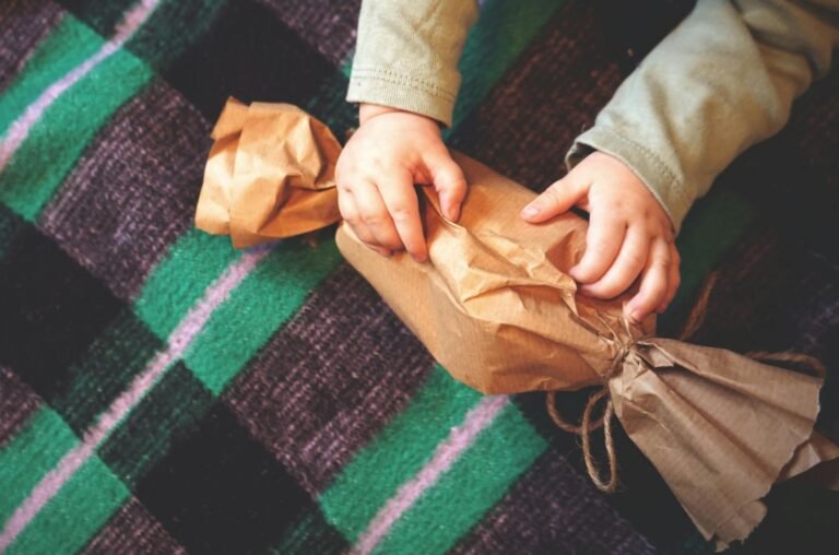 A child unwrapping a brown paper gift on a colorful checkered blanket.